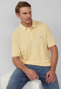 Jasnożółta koszulka polo o teksturowanym wykończeniu, krótkie rękawy i mała łatka z logo po lewej stronie klatki piersiowej, zestawiona z niebieskimi dżinsami.