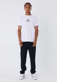 T-shirt bianca in cotone con logo nero, abbinata a pantaloni neri larghi e sneaker grigie, su uno sfondo neutro.