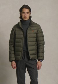 Casaco acolchoado verde-oliva com costuras horizontais, fecho de correr e um pequeno logótipo. Usado sobre uma camisola de gola alta escura e calças cinzentas.