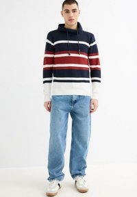 Joven de pie con una sudadera con capucha de punto a rayas en azul marino, rojo y blanco, vaqueros azul claro y zapatillas blancas con suelas de goma.