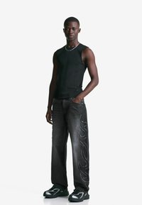 Giovane uomo in piedi, indossa una maglietta nera senza maniche, jeans neri con design di fiamme su un lato e sneaker nere, su uno sfondo bianco uniforme.