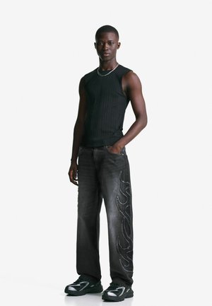 Young man standing, wearing a black sleeveless top, black jeans with flame designs on one side, and black sneakers, against a plain white background.