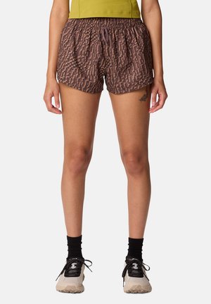 Femme portant un short marron à motifs, des chaussettes noires, des baskets beiges et noires, et un top court jaune sur un fond uni.
