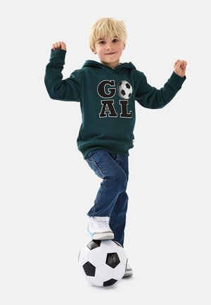 Grüne Kapuzenjacke mit dem Text "GOAL" und einem Fußball-Design, kombiniert mit blauen Jeans und weißen Turnschuhen, die auf einem schwarz-weißen Fußball stehen.
