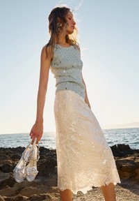 Young woman holding silver sandals, wearing a light blue sleeveless top and a white lace skirt, standing on rocky shore by the sea at sunset.