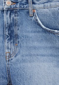 Gros plan sur la poche d'un jean en denim bleu clair avec des rivets métalliques, une couture orange et une texture délavée près de la taille avant.