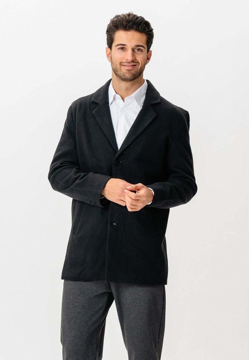 Mand med skæg og kort hår iført en sort frakke, hvid skjorte og grå bukser, står og smiler mod en hvid baggrund.