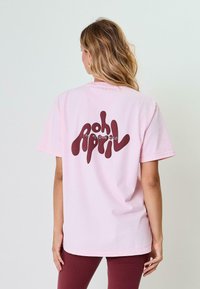 Růžové bavlněné tričko s vlnitým tmavě vínovým grafickým textovým designem na zadní straně. Volný střih s kulatým výstřihem a krátkými rukávy.