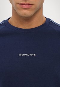 Námornícky modrá mikina s okrúhlym výstrihom, so bielym logom "MICHAEL KORS" umiestneným na strede a textúrovaným detailom látky pozdĺž ramien.