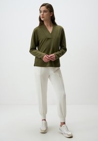 Maglia lunga verde oliva con scollatura a V e spacchi laterali, abbinata a pantaloni bianchi a sigaretta e sneakers beige con dettagli testurizzati.