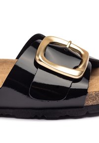 Sandale en cuir verni noir avec une boucle carrée dorée, une semelle intérieure en liège et une semelle extérieure en caoutchouc noir texturé. Surface lisse avec une finition brillante.