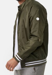 Veste bomber vert olive avec fermeture éclair, boutons-pression blancs, et poignets rayés blancs et verts olive ; texture lisse avec poches latérales.