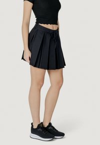 Gonna nera plissettata con una vita aderente e un dettaglio a fiocco, abbinata a scarpe sportive nere con suola bianca e tomaia texturizzata.