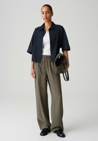 Chemise noire à manches courtes sur un t-shirt blanc, assortie à un pantalon ample vert olive et des chaussures noires à enfiler, tenant un sac à textures sombre.