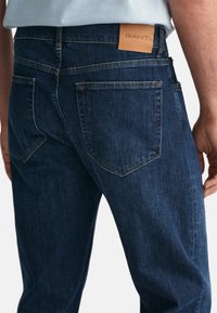 Jeans din denim de culoare albastru închis cu un design ajustat, având un etichetă din piele GANT la talie și două buzunare pe spate cu cusături curbe.