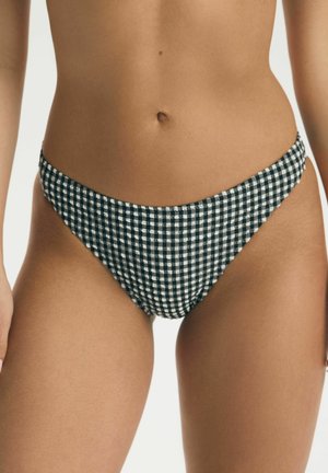 Žena v čiernych a bielych kockovaných spodných dieloch bikín, ukazujúca stred tela a horné stehná na jednofarebnom pozadí.