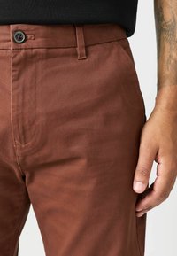 Braune Baumwollhosen mit glatter Textur, ausgestattet mit einer Vordertasche und einem einzelnen Knopfverschluss, teilweise von einer Hand gehalten.