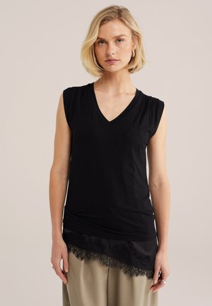 Heledate juustega naine, kes kannab musta varrukateta V-kaelusega topi pitsist äärega ja beežeid pükse, seisab ühevärvilise hele tausta ees.