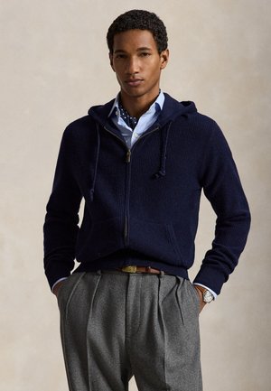 Homme portant un sweat à capuche bleu marine zippé sur une chemise bleue avec un foulard à pois autour du cou et un pantalon plissé gris avec ceinture marron.