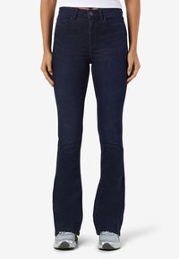 Personne portant un jean flare bleu foncé, un top blanc court et des baskets argentées, debout sur un fond clair.