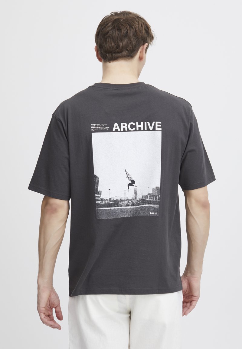 Grå bomulds T-shirt med korte ærmer, med et hvidt grafisk tryk på ryggen og teksten "ARCHIVE." Struktureret stof.