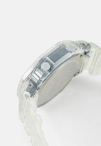 Montre en plastique transparent avec un boîtier arrière en acier argenté, deux boutons gris clair et un bracelet segmenté. Caractéristiques d'un design clair et angulaire.