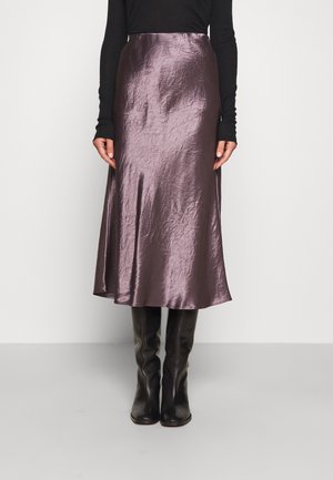 Kobieta w błyszczącej sukience midi w kolorze mauve, czarnym bluzce z długim rękawem i czarnych, sięgających do kolan butach, stojąca na tle jasnego jednolitego tła.