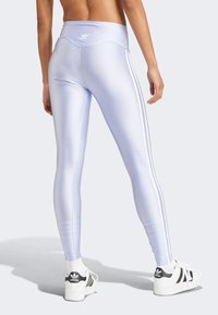 Person som bär glänsande ljuslila Adidas leggings med tre vita ränder på sidan och vita Adidas sneakers som står.