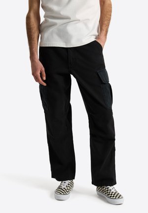 Man draagt zwarte cargobroek met klepzakken, een wit T-shirt en zwart-witte geruite sneakers, staand met één hand in de zak.