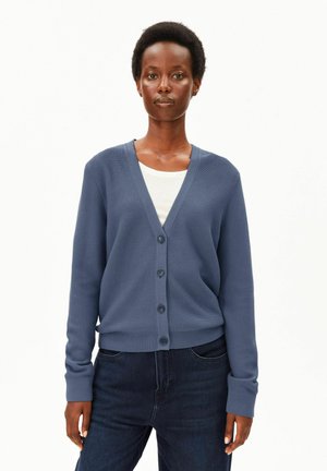 Frau trägt eine blaue, geknöpfte Strickjacke über einem weißen Top und dunklen Jeans und steht vor einem schlichten hellen Hintergrund.