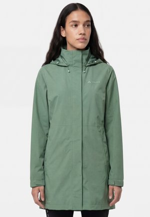 Vrouw draagt een lange groene waterdichte jas met capuchon, ritssluiting aan de voorkant en een logo op de linkerborst, staand tegen een effen achtergrond.