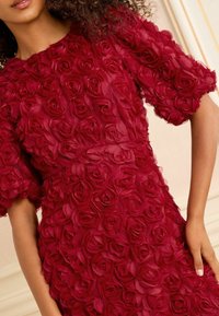 Red dress made of textured fabric adorned with three-dimensional rose motifs, featuring short puff sleeves and a fitted waist.