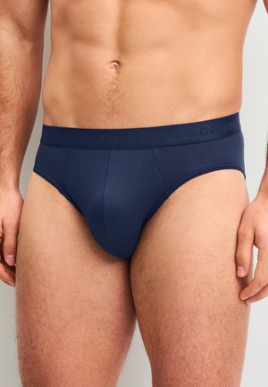 Männlicher Oberkörper und obere Beine, die dunkelblaue Slips mit gebrandetem Bund vor einfarbigem Hintergrund tragen.