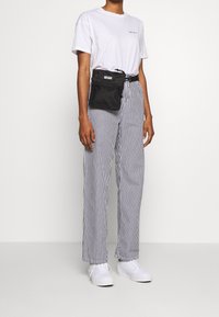 Czarna nylonowa nerka z zamkiem i regulowanym paskiem noszona na białej koszulce oraz szerokich spodniach w paski, zestawiona z białymi trampkami.