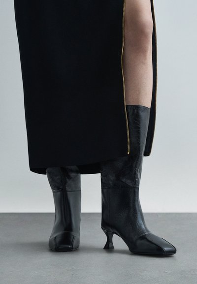 Fekete bőr, középmagas csizmák négyzetes orral és vékony sarokkal, fekete szoknyával párosítva, amelyen egy arany cipzáros hasíték található, felfedve a meztelen lábat.