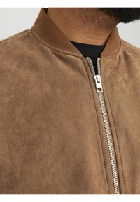 Gros plan d'une personne portant une veste en daim marron avec une fermeture éclair argentée et un col côtelé sur une chemise noire.