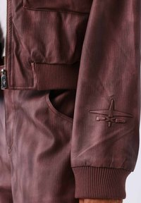 Bordeaux bomberjack met een gestructureerde afwerking, geribde manchetten, voorzien van een logopatch op de mouw en een zijzak. Ritsafsluiting.