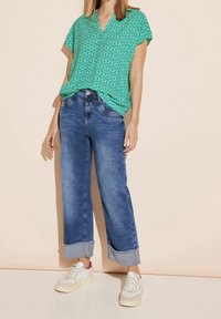 Zielona bluzka w wzory z dekoltem w serek, połączona z szerokimi niebieskimi dżinsami z rolowanym dołem i białymi trampkami.