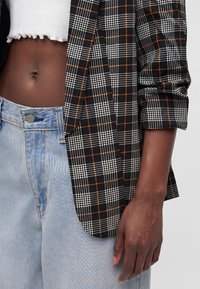 Personne à la peau foncée portant un jean bleu clair, un top court blanc et un blazer à carreaux noir avec des lignes orange et blanches, bras détendu le long du corps.