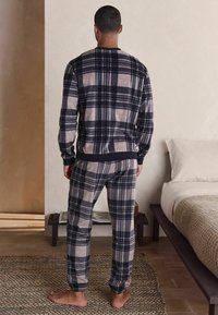 Mann, der barfuß auf einem Teppich im Schlafzimmer steht, trägt ein passendes Oberteil und eine Hose aus kariertem Schwarz und Beige mit langen Ärmeln, und schaut weg.