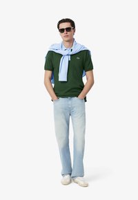 Grön polo skjorta, ljusblå tröja draperad över axlarna, ljusblå jeans och vita sneakers. Modell som bär solglasögon.