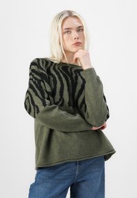 Grüner Strickpullover mit schwarzem Zickzackmuster, entspannte Passform, runder Ausschnitt und gerippte Bündchen. Kombiniert mit blauen Jeans.