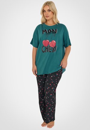 Lyshåret kvinde stående barfodet iført en teal oversized T-shirt med kirsebær og teksten "Mon Cheri" samt sorte bukser med farverige kirsebær- og hjerte-mønstre.
