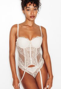 Corsetto in pizzo beige con bordi smerlati e pannelli trasparenti, dotato di spalline regolabili e chiusura frontale. Include bretelle reggicalze.