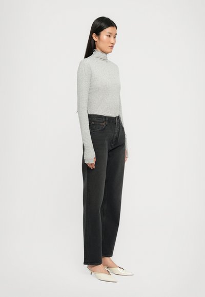 Eine Frau steht im Profil und trägt einen hellgrauen Rollkragenpullover, schwarze hochgeschnittene Jeans und cremefarbene spitze Absatzschuhe vor einem schlichten Hintergrund.