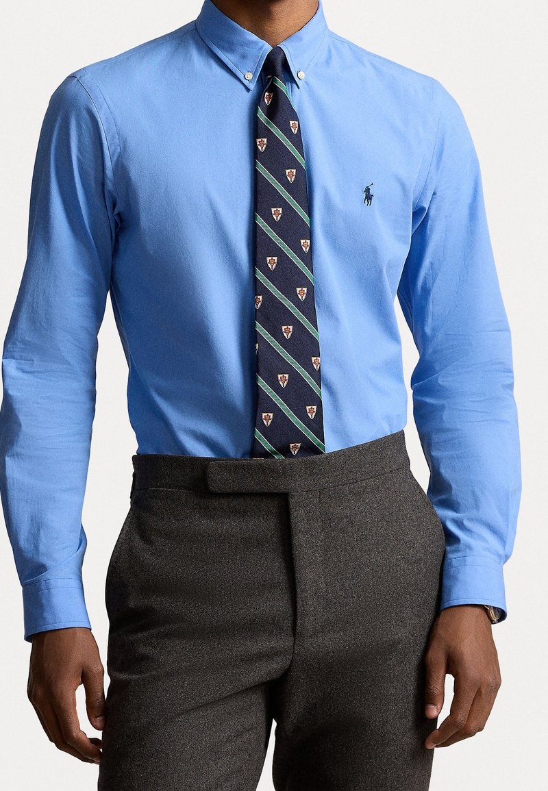 Uomo che indossa una camicia blu con bottoni, una cravatta blu navy a motivo e pantaloni grigi scuri a vita alta con chiusura laterale a linguetta.