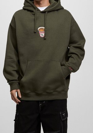 Personne portant un sweat à capuche vert olive avec un motif de tasse de café rose et un pantalon cargo noir avec des coutures blanches.