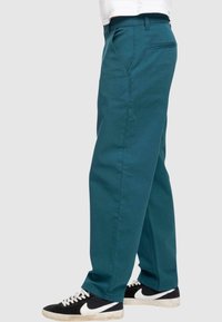 Pantaloni in cotone verde acqua con design a gamba dritta, tasche laterali, una tasca posteriore e indossati con sneakers nere.