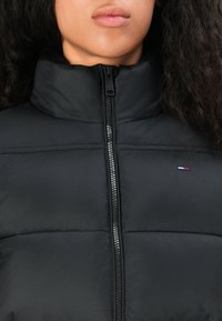 Sort pufferjakke med høj krave, lynlås lukning og diskret horisontal syning. Har et lille logo med røde, hvide og blå accenter.