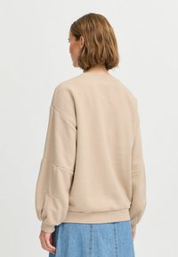 Sweatshirt beige avec des manches bouffantes, col rond et texture douce. Présente une taille ajustée et une silhouette décontractée. Associé à une jupe en jean.